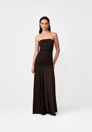 Žena v čiernych šatách bez ramienok s riasením a širokými nohavicami, stojaca pred jednoduchým bielym pozadím, nosí zlaté kruhové náušnice.