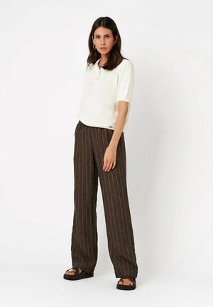 Femme aux longs cheveux foncés portant un top blanc à manches courtes, un pantalon large marron à fines rayures et des sandales noires, debout sur fond blanc.