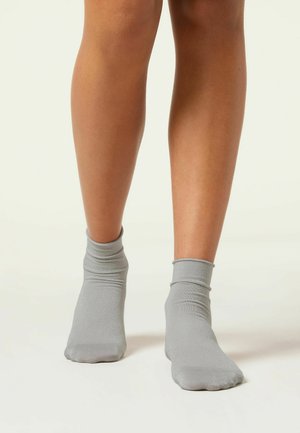Grå ankelsockor i lätt, texturerat tyg med en följsam mudd, vilande på bara ben mot en ljus bakgrund.