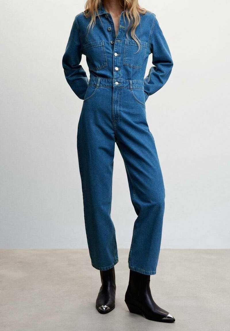 Femme portant une combinaison en denim bleu à manches longues et des bottines noires avec embouts métalliques, debout devant un fond uni.