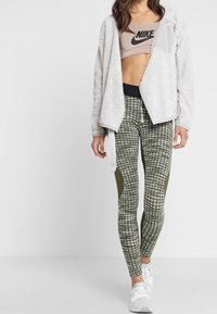 Kobieta w beżowej, krótkiej bluzie Nike, białej polarowej kurtce, wzorzystych zielonych legginsach i białych butach do biegania, stojąca na jednolitym tle.