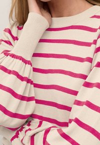 Pull en tricot avec une base crème et des rayures horizontales roses vives. Présente des manches bouffantes et des poignets côtelés pour un supplément de texture et de détail.