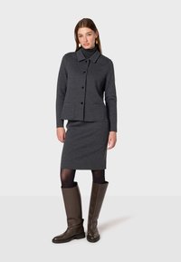 Veste en mélange de laine gris foncé avec un col, un devant à boutons et deux poches, associée à une jupe crayon ajustée. Bottes brunes jusqu'aux genoux.