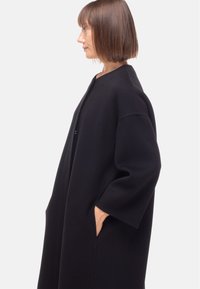 Cappotto lungo nero, oversize, con maniche larghe, tasche laterali e design minimalista. Realizzato in tessuto testurizzato con una finitura liscia.