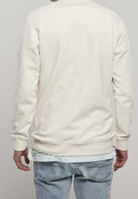 Sudadera color crema con una textura suave, con puños y dobladillo acanalados. Llevada sobre una camisa de color azul claro, combinada con jeans de mezclilla claros.