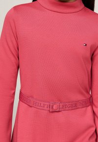 Sukienka z długim rękawem w jasnoróżowym kolorze, z wysokim kołnierzem i paskiem w talii z napisem "HILFIGER" w fioletowej literze. Gładka tekstura.