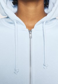 Ljusblå zip-up hoodie med en mjuk textur, med en silverfärgad dragkedja och justerbara snoddar på huvan.