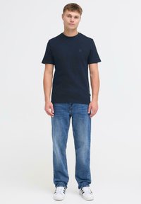 Tamsiai mėlyna trumpų rankovių marškinėlių su tekstūruota medžiaga derinys su šviesiai mėlynomis tiesiosios kojos džinsomis ir baltais sportbačiais su juodomis juostelėmis.