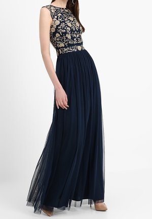 Kvinde iført en ærmeløs marineblå kjole med gyldent blomstermønster på overdelen og et flydende tylskørt, kombineret med beige hæle.