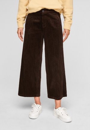 Persona con pantalones de pana de pierna ancha de color marrón oscuro y zapatillas blancas, con las manos relajadas a los lados.
