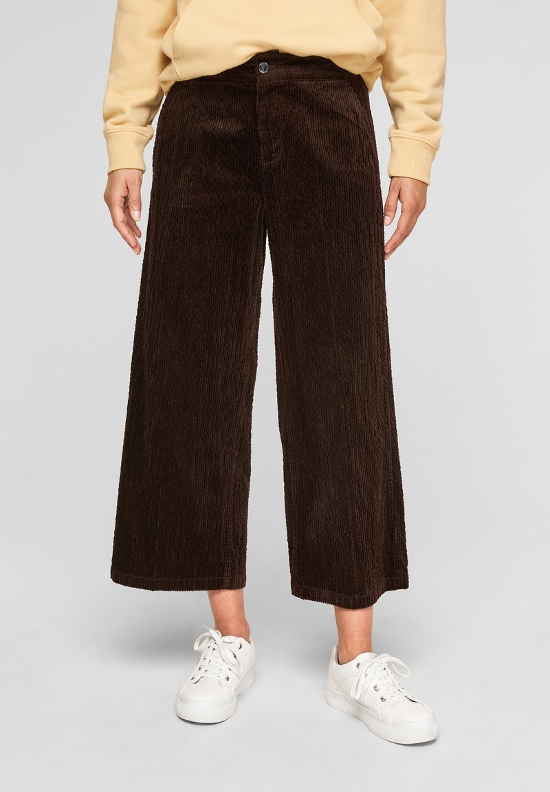 Personne portant un pantalon large en velours côtelé marron foncé et des baskets blanches, les mains détendues le long du corps.