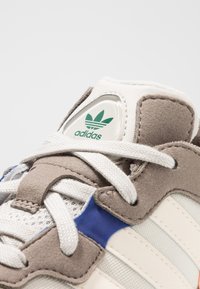 Beige och grå sneaker med ett meshdesign, som har en vit lädersnörning och blå detaljer, samt pryds av en grön "adidas"-logotyp.