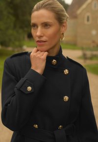 Tamno mornarsko plavi vuneni kaput s dvostrukim kopčanjem, zlatnim dugmadi, epauletama na ramenima i odgovarajućim remenom. Glatka tekstura.