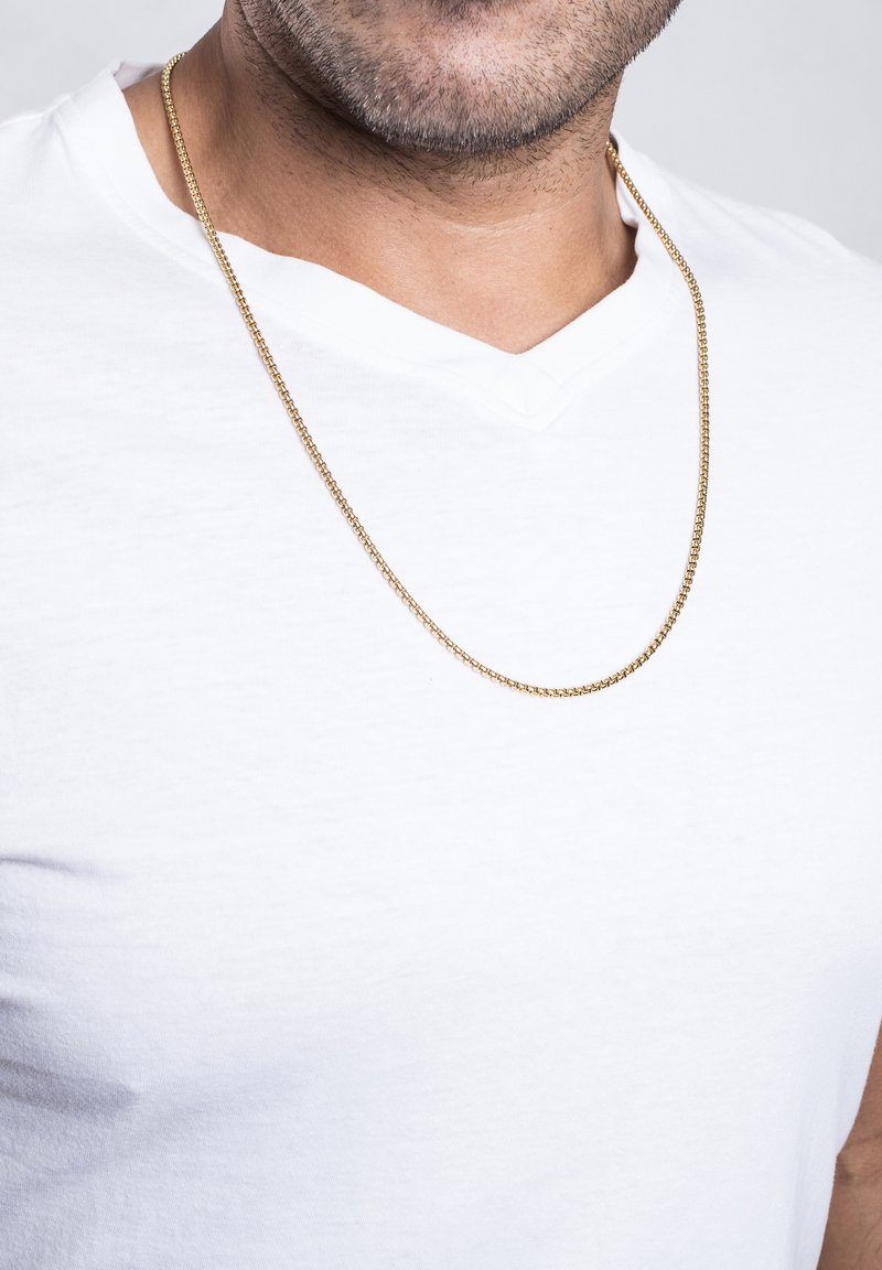 Collar de cadena de oro reposando sobre una camiseta blanca con cuello en forma de V, presentando un diseño liso y redondeado con eslabones entrelazados y un brillo sutil.