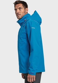 Blauwe waterdichte jas met capuchon, voorzien van verstelbare koordjes, zijzakken en een logo op de linkerschouder. Soepele textuur.