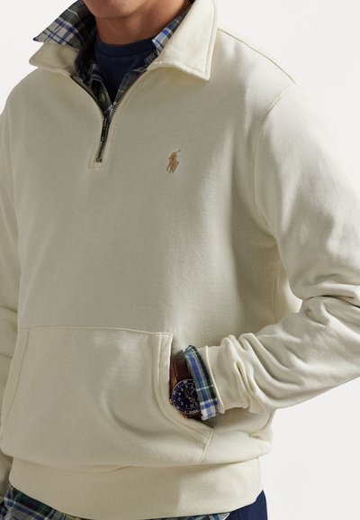 Kremfarget genser med kvart-zip design, som har et strukturert fleece-materiale, brystlogo og en frontlomme. Under er en rutete skjorte synlig.