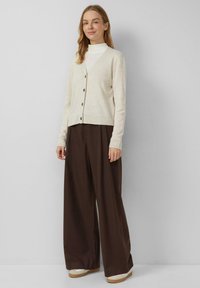 Beżowy kardigan z dekoltem w serek, z guzikami z przodu i ściągaczami na rękawach, noszony na białym golfie i brązowych spodniach o szerokich nogawkach, z casualowym obuwiem.