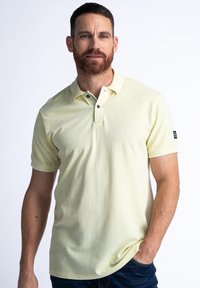 Jasnożółta koszulka polo z krótkim rękawem, żebrowanym kołnierzykiem i zapięciem na trzy guziki. Posiada małe czarne logo na lewym rękawie.