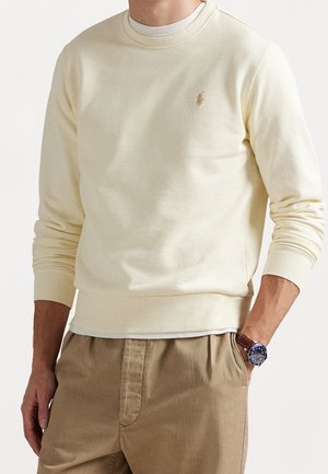 Man met een crèmekleurige trui met lange mouwen en een klein geborduurd logo, beige broek en een horloge met een blauwe wijzerplaat en een bruine leren band.