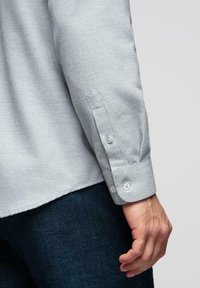 Poignet de chemise à manches longues gris clair avec deux boutons, portée par une personne à côté d'un jean bleu foncé, main détendue le long du corps.