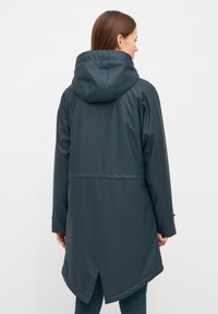 Cappotto da pioggia lungo e verde scuro con cappuccio, che presenta una vita svasata, tasche laterali e un orlo leggermente curvo. Materiale liscio e impermeabile.