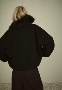 Crni oversized sako s visokim ovratnikom od umjetnog krzna, s teksturiranim materijalom i spuštenim ramenima, u kombinaciji s tamnim hlačama.