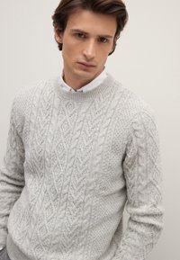 Hellgrau gestrickter Pullover mit strukturierten Mustern, Rundhalsausschnitt und gerippten Bündchen. Über einem weißen Hemd mit Kragen getragen.