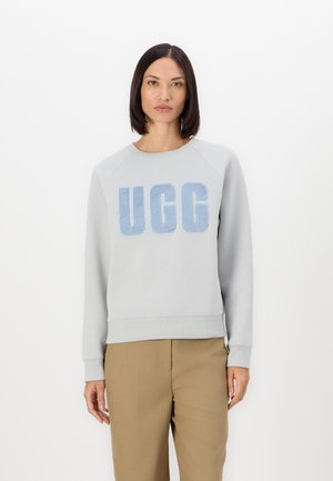 Felpa grigia chiara con logo "UGG" in pile blu. Presenta maniche raglan e orlo a coste. Indossata con pantaloni beige, il modello si trova davanti a uno sfondo bianco.