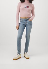 Suéter de cuello alto de canalé rosa con logotipo bordado, combinado con vaqueros ajustados de color azul claro y zapatillas blancas con detalles amarillos.