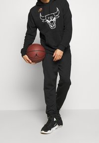 Person i svart Chicago Bulls-huvtröja och joggingsbyxor som håller en basketboll, stående mot en enkel vit bakgrund.