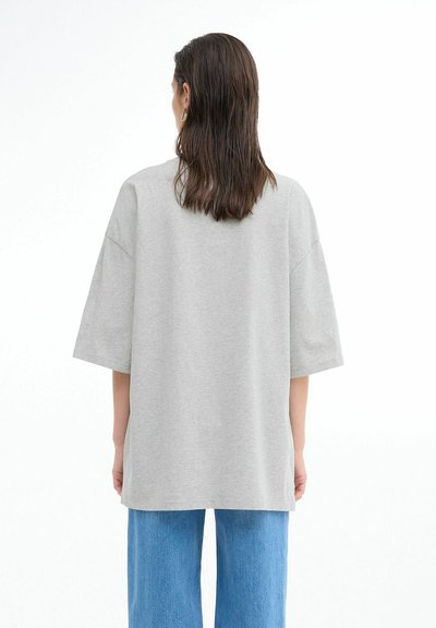 Sivá oversized tričko s prehnutými ramenami, vyrobené z mäkkej tkaniny, s okrúhlym výstrihom a voľným strihom. Nosené s svetlomodrými džínami.
