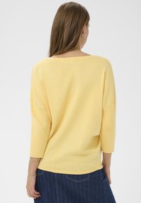 Pull léger jaune avec un large décolleté et des manches trois-quarts. Texture en maille douce et détail de couture latérale. Porté avec une jupe en denim.
