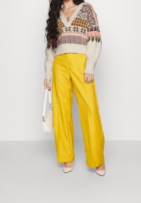 Stickad tröja med flerfärgade mönster, V-ringning; vida gula byxor; håller en vit handväska; neutral bakgrund.