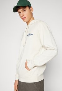 Felpa color crema con tasca a marsupio e logo blu. Indossata con pantaloni grigi e un cappellino verde. Tessuto in cotone morbido e casual.