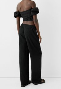 Haut en tissu noir texturé avec manches ballon, assorti à un pantalon noir ample. La tenue comprend une ceinture élastique et des sandales décontractées.