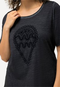Černý top s kulatým výstřihem a krátkými rukávy. Vyznačuje se výrazným embosovaným vzorem ve tvaru srdce s květinovými motivy a zakřivenými liniemi.