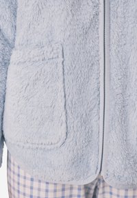 Helleblaue, fuzzy Fleecejacke mit weicher Textur, ausgestattet mit zwei Vorderaschen und einem Reißverschluss, der mit einem kontrastierenden Besatz umrahmt ist.