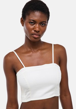 Jeune femme noire aux cheveux courts portant un crop top blanc sans manches sur un fond clair uni.