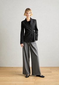 Veste noire cintrée avec un détail de ceinture, associée à un pantalon large gris et des ballerines noires, le tout sur un fond neutre.