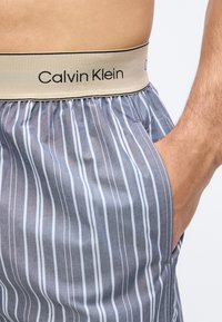 Ανδρικός κορμός που φορά γκρι και άσπρα κάθετα ριγέ εσώρουχα μποξέρ Calvin Klein με μπεζ ελαστική μέση και χέρι στην πλευρική τσέπη.