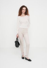 Λευκό μακρυμάνικο τοπ από δαντέλα με κυματιστό τελείωμα, συνδυασμένο με ανοιχτό μπεζ ψηλόμεσο παντελόνι. Το μοντέλο κρατά μια μικρή μαύρη τσάντα χειρός και φορά μαύρα φλατ παπούτσια.