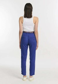 Blauwe broek van een lichtgewicht stof, met een relaxte pasvorm, zijzakken en een gemusterde riem. G gedragen met een witte tanktop en sneakers.