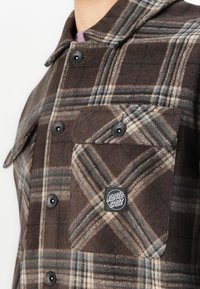 Bruine en grijze geruite button-up shirt met zwarte knopen en een klein vierkant Santa Cruz logo patch op de borstzak.