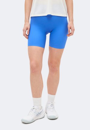 Persoon draagt felblauwe geribbelde biker shorts, witte sportschoenen, sokken en een wit topje, staand voor een effen achtergrond.