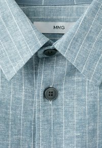 Chemise habillée bleu clair avec des rayures verticales blanches, un col pointu et des boutons noirs, montrant l'étiquette de la marque à l'intérieur du col.