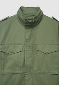 Giacca verde oliva con collo alla coreana, due grandi tasche con patta e una texture liscia e opaca. Presenta spalline sulle spalle e un taglio dritto.