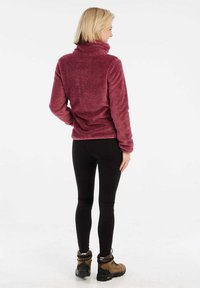 Pullover in pile color bordeaux con collo alto, superficie strutturata e vestibilità morbida, abbinato a leggings neri e scarponi da trekking marroni.