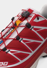 Κόκκινο αθλητικό παπούτσι με λευκά και κίτρινα στοιχεία, διαθέτει σύστημα δέσιμου με γρήγορο τράβηγμα με κόκκινους ελαστικούς κορδόνια και μαύρα πλαστικά κλειδώματα.