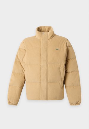 Homme portant une veste matelassée beige, un pull rose, un jean bleu et des baskets blanches, debout les mains dans les poches de la veste.
