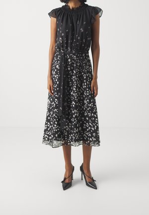 Femme portant une robe midi noire avec des taches abstraites blanches, un col volanté, des manches courtes, une taille nouée et des talons hauts noirs pointus.